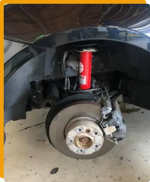 Brake Repair In Tomball TX At Jon’s Auto & Diesel Repair. Close-up of a car’s wheel well showing the suspension, red shock absorber, brake disc, and caliper, with the tire removed and the car elevated.