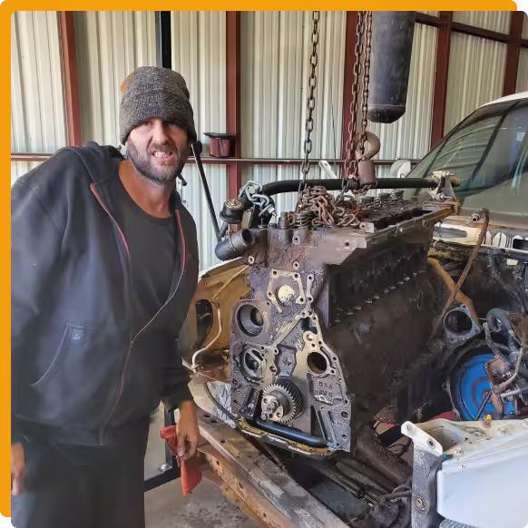 Auto & Diesel Repair In Tomball TX At Jon’s Auto & Diesel Repair. A man in a beanie and hoodie stands next to a car with its engine exposed inside a garage, smiling at the camera.