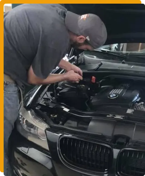 Auto Diagnostics & electrical repair In Tomball TX At Jon’s Auto & Diesel Repair. A man wearing a cap and gray shirt is working under the hood of a black BMW car, appearing to repair or inspect the engine.