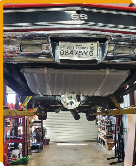Exhaust System Repair In Tomball TX At Jon’s Auto & Diesel Repair. A classic black car with an SS emblem is lifted in a garage. Tools, car parts, and shelves surround the car. The view shows the undercarriage and an expired license plate.