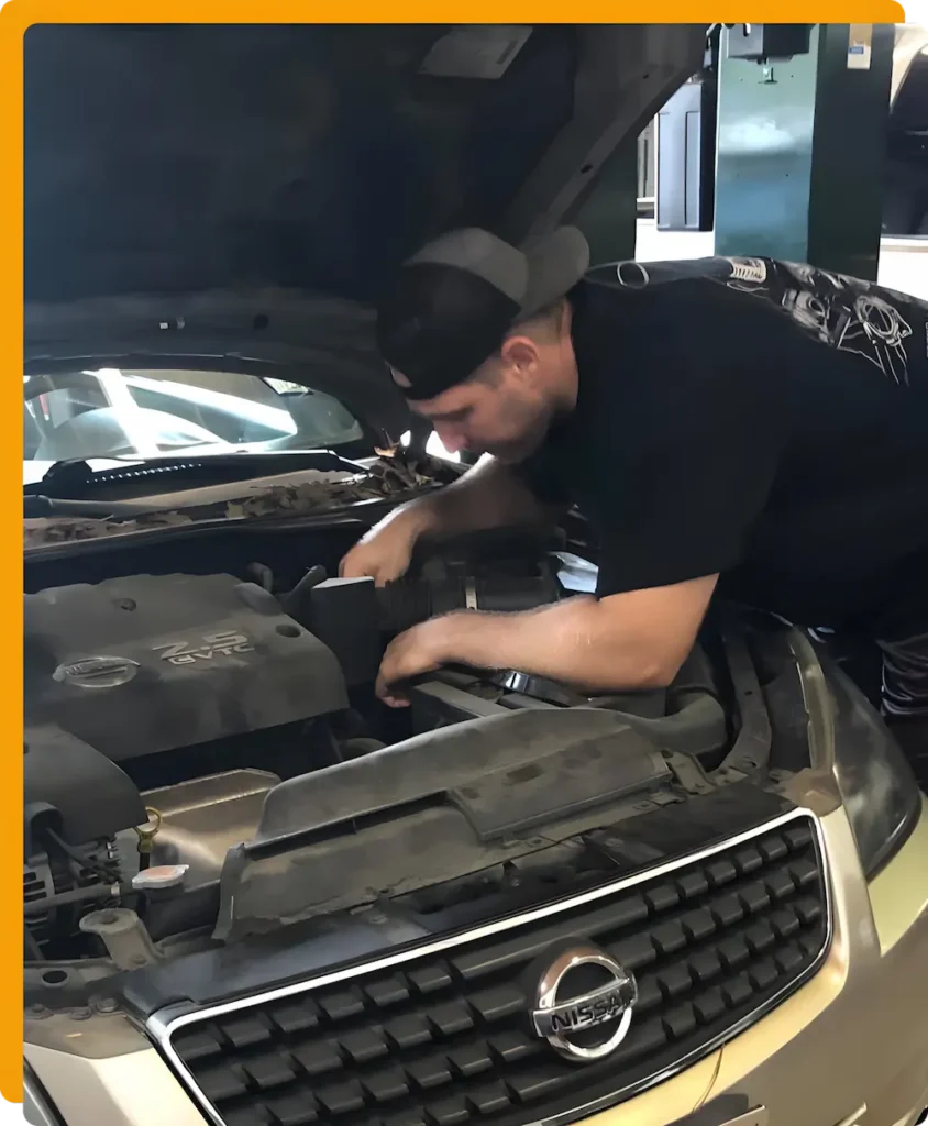 Auto & Diesel Repair Mechanic In Tomball TX At Jon’s Auto & Diesel Repair. A man wearing a backwards cap works under the hood of a Nissan car in a garage, performing maintenance or repairs on the engine.